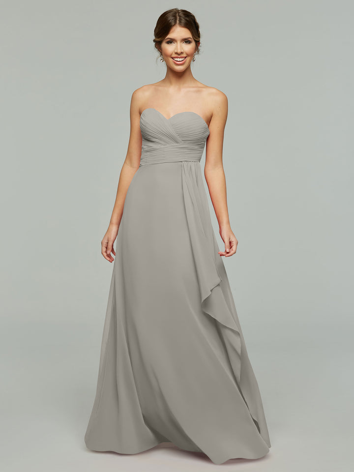 A woman smiles against a light gray background wearing Avery Austin style 80046, a timeless strapless chiffon gown with a floor-length light gray skirt, sweetheart neckline, and draped ruched bodice.