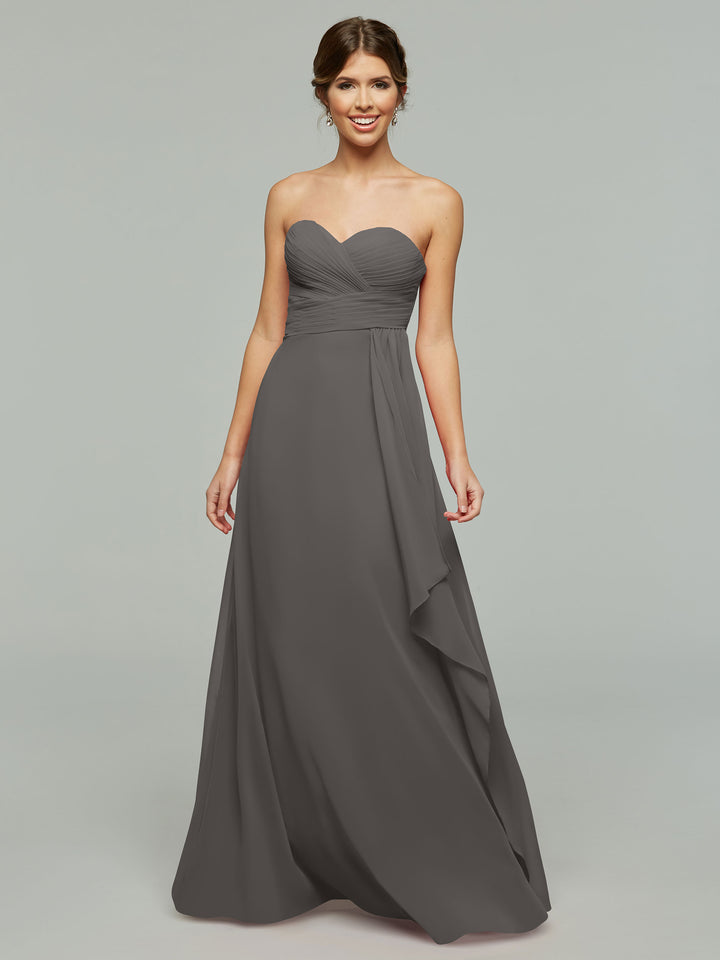 A woman smiles in front of a plain gray background, wearing Avery Austin’s 80046—a timeless strapless chiffon dress in charcoal gray with a sweetheart neckline and ruched bodice. Her hair is styled in a low, loose updo.
