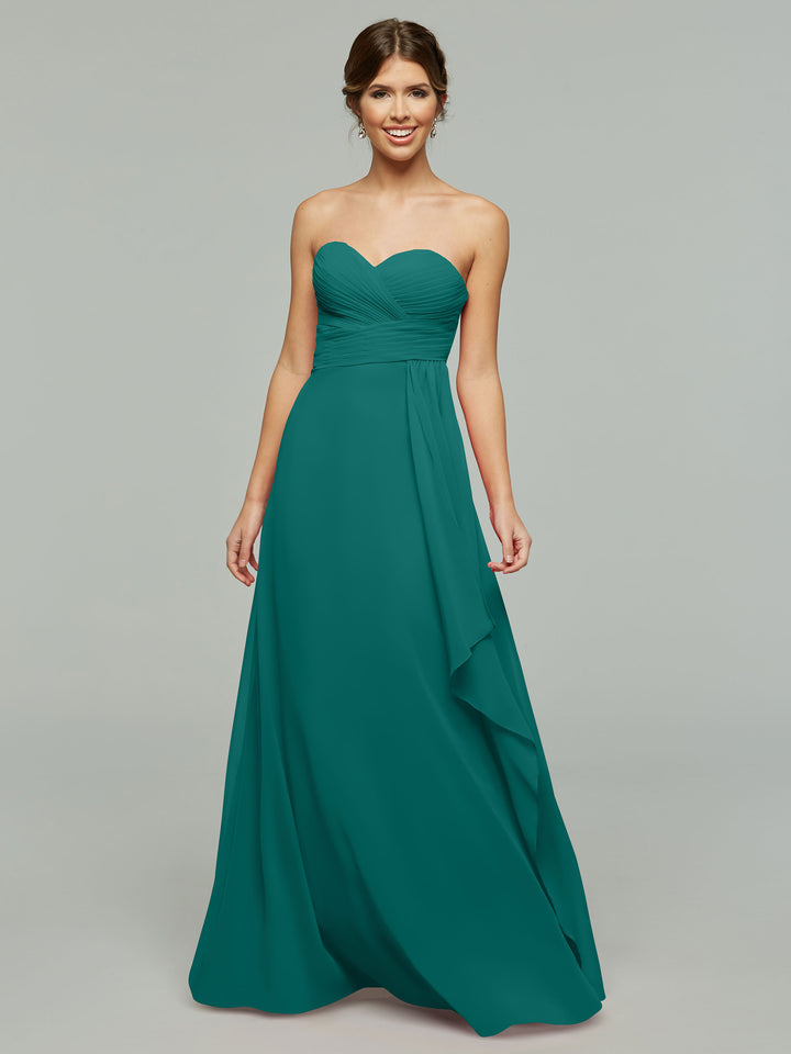 A woman smiles against a gray background, wearing Avery Austin’s timeless strapless chiffon dress (style 80046) in teal, featuring a sweetheart neckline and ruched bodice, with her hands relaxed by her sides.
