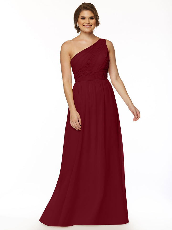 A woman with short brown hair and earrings smiles against a white background in Avery Austin’s 80047: a floor-length, one-shoulder chiffon dress with a ruched bodice.