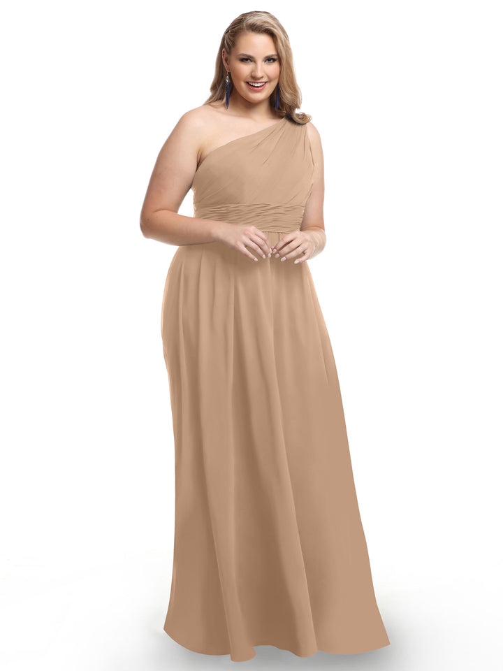 A woman with long, light brown hair smiles in the Avery Austin 80047, a buff floor-length one-shoulder chiffon dress with a modern ruched bodice, standing against a plain white background.