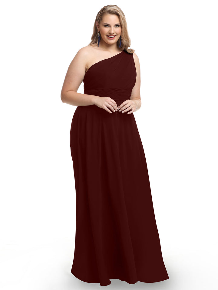 A woman smiles against a white background, modeling Avery Austin’s 80047 sleeveless, one-shoulder chiffon gown in merlot with a modern twist and floor-length design.