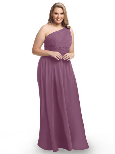 A woman stands smiling in Avery Austin’s 80047, a floor-length mauve one-shoulder chiffon gown with a ruched bodice, set against a plain white background for a fresh take on classic elegance.