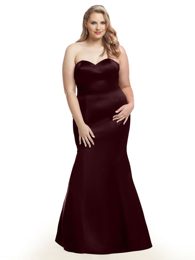 A woman with long, wavy blonde hair models Avery Austin 80072, a strapless sweetheart fit and flare dress in Bordeaux, standing against a white background with a slight smile and one hand on her hip.