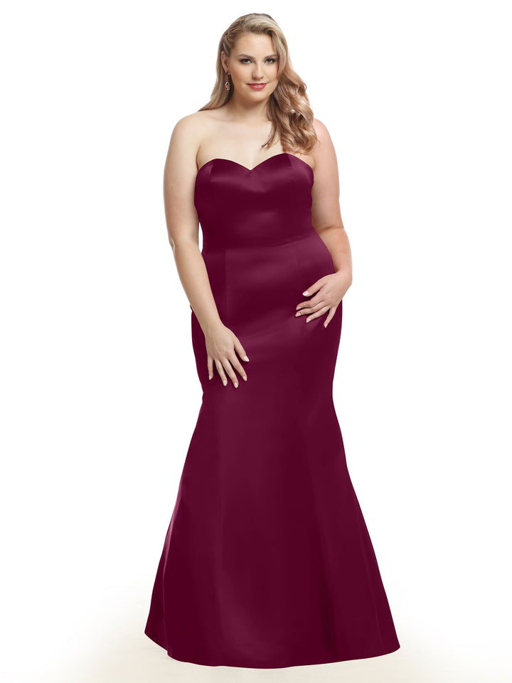 A woman models Avery Austin style 80072, a plum satin floor-length strapless mermaid bridesmaid dress, standing against a white background with one hand on her hip.