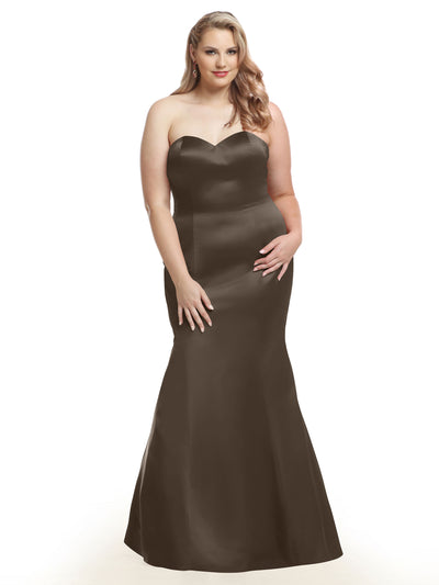 A woman with long, wavy blonde hair models the Avery Austin 80072 gown in taupe, featuring a strapless sweetheart neckline and fitted, floor-length silhouette. She stands against a white background, smiling gently with one hand on her hip.