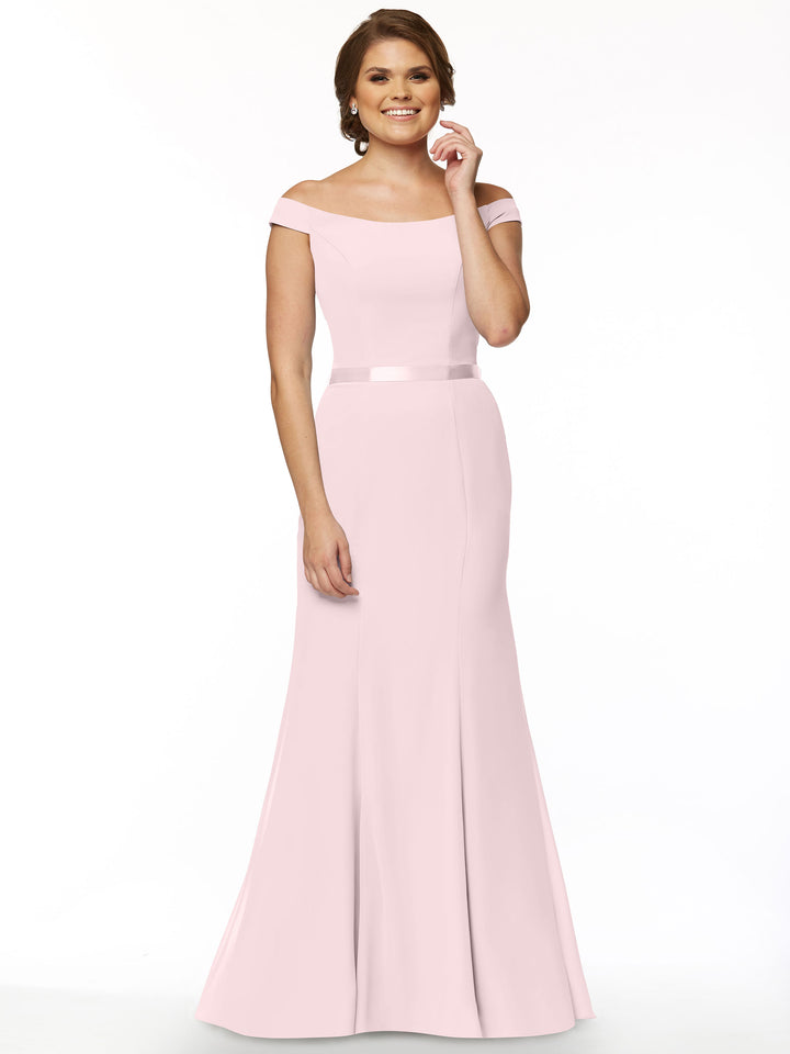A woman smiles gracefully in Avery Austin’s floor-length, off-the-shoulder bridesmaid dress (Style 80079) in pale pink, featuring a fitted bodice and slight flare, posed against a plain white background.