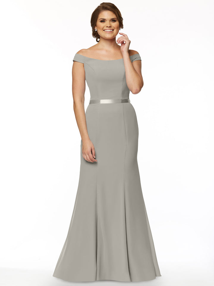 A woman models Avery Austin’s 80079, a floor-length, off-the-shoulder bridesmaid dress in light gray with a satin waistband, smiling with one hand on her hip against a plain white background.