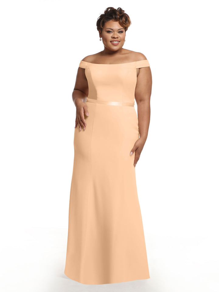 A woman stands against a plain white background, smiling with one hand on her hip, wearing the Avery Austin 80079 blush off-the-shoulder floor-length fit-and-flare bridesmaid dress.