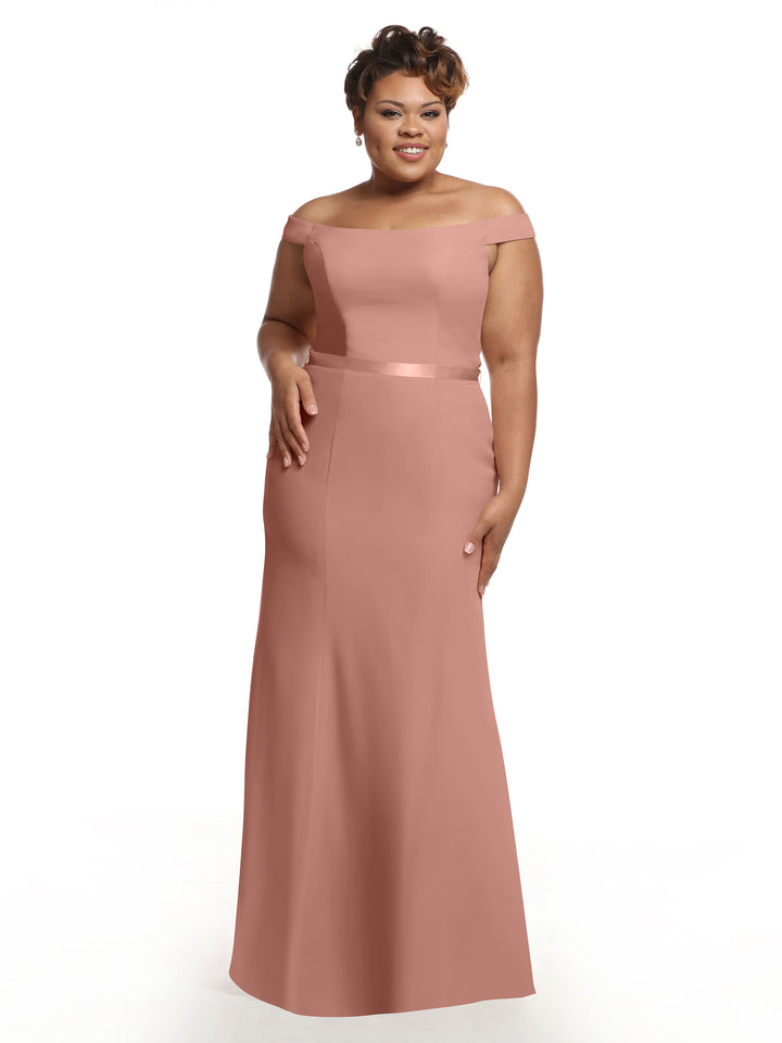 A woman smiles gently while modeling Avery Austin’s 80079: a dusty rose, floor-length, off-the-shoulder bridesmaid dress with a fitted bodice and satin waistband, standing against a plain white background.