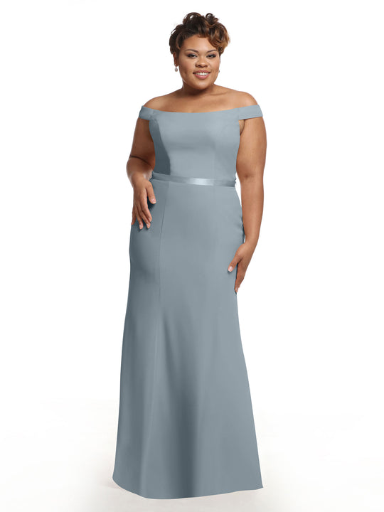 A woman stands and smiles against a white background, wearing Avery Austin’s light blue off-the-shoulder bridesmaid dress (style 80079) featuring a satin waistband.