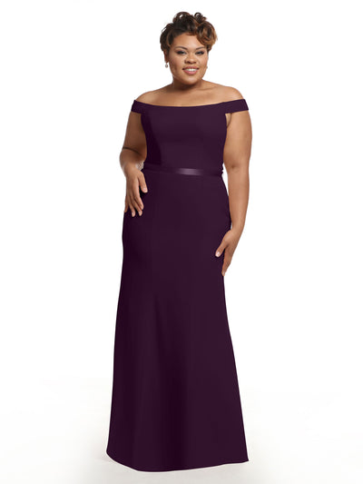 A woman models Avery Austin’s floor-length, off-the-shoulder bridesmaid dress (Style 80079) in deep purple, standing against a white background with one hand at her side and the other on her waist, smiling.