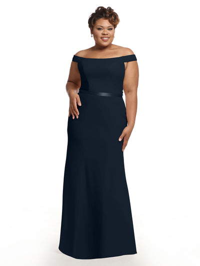 A woman models Avery Austin style 80079, an off-the-shoulder, floor-length navy blue chiffon bridesmaid dress, standing against a white background with a smile, one hand at her side and the other resting on her hip.