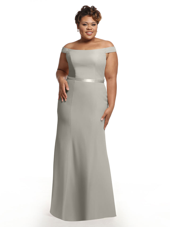 A woman smiles against a white background wearing Avery Austin’s style 80079, a light gray off-the-shoulder bridesmaid dress with a satin waistband.