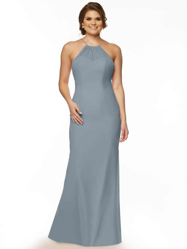 A woman stands smiling in Avery Austin’s 80083 gown—a sleeveless, floor-length chiffon dress in dusty blue with a halter neckline and chic keyhole back—set against a plain white background.