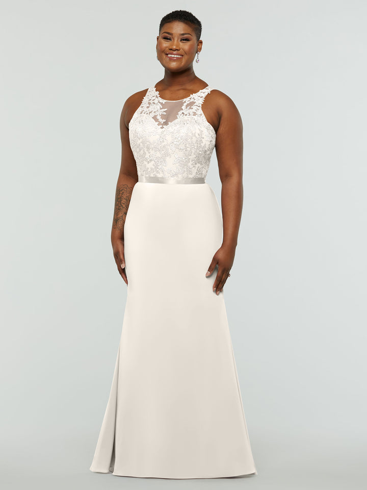 A person smiles in Avery Austin’s 80095: an ivory, sleeveless, floor-length gown with a fitted illusion lace bodice and a smooth skirt. The background is plain and light-colored.