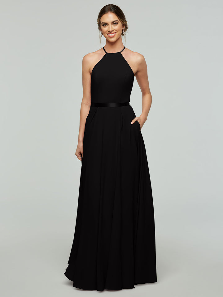 A woman is smiling in the Avery Austin 80099 sleeveless floor-length A-line dress with a halter neckline, satin ribbon belt, and her hair in a low updo. She stands with one hand in her pocket against a light gray background.