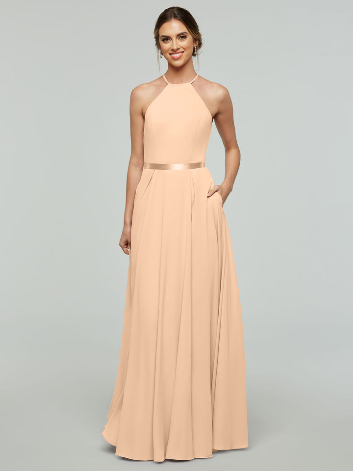 A woman wearing Avery Austin’s dress 80099—a long, sleeveless light peach A-line with a halter neckline and satin ribbon belt—smiles with one hand in her pocket against a plain light gray background.