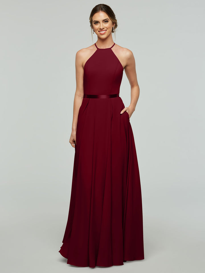 A woman wearing Avery Austin 80099—a sleeveless, floor-length burgundy A-line dress with a halter neckline and fitted waist—smiles with her hair tied back, hand in pocket, standing against a plain light background.