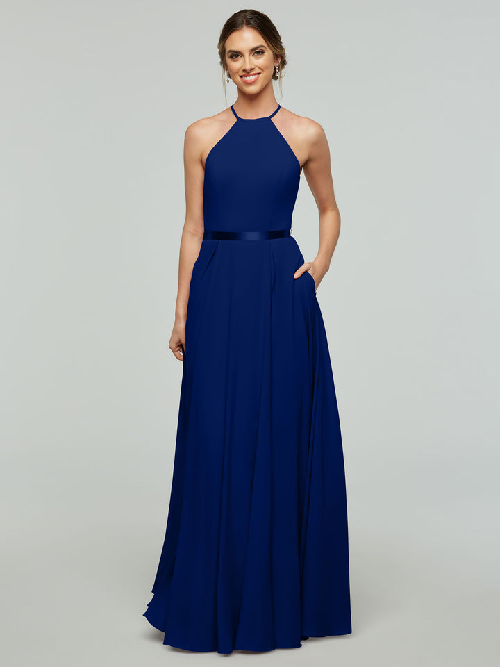 A woman smiles in Avery Austin style 80099, a sleeveless, floor-length royal blue A-line dress with a halter neckline, fitted bodice, satin ribbon belt, and pockets. Her hair is styled in an updo against a plain background.