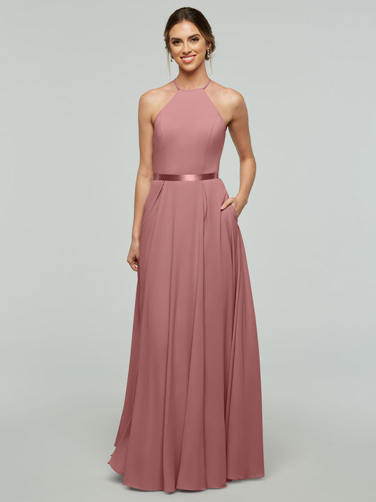 A woman models Avery Austin style 80099, a dusty rose sleeveless A-line gown with halter neckline, satin waistband, and pockets, standing against a plain light gray background with her hair in a low updo and smiling.