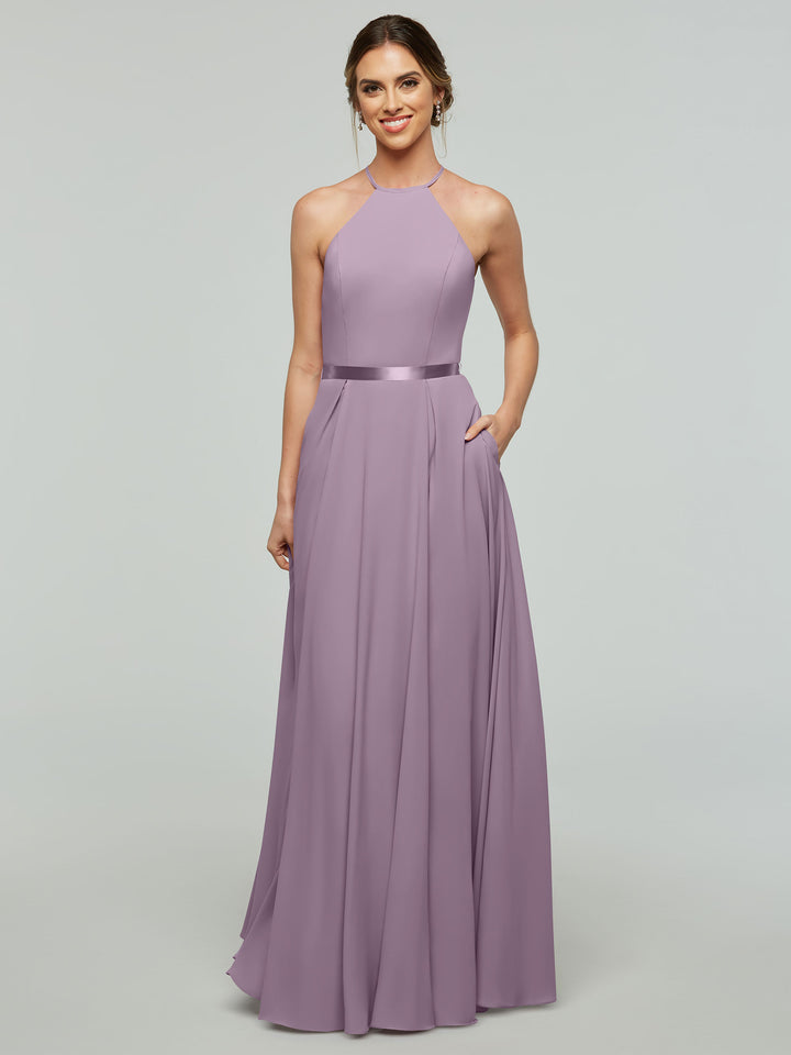 A smiling woman models the Avery Austin 80099 dress—a lavender, sleeveless A-line gown with a halter neckline and satin ribbon belt—posing with one hand in her pocket against a light gray background.