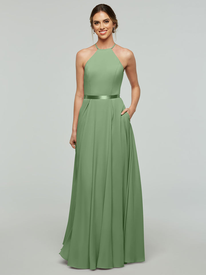A woman stands smiling with one hand in her pocket, modeling the Avery Austin 80099: an A-line, floor-length sage green gown featuring a halter neckline and satin ribbon belt, set against a plain light background.