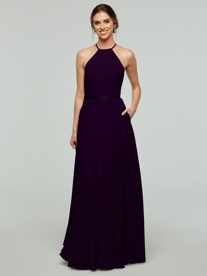 A woman stands against a light gray background, smiling with her hair pulled back, wearing the Avery Austin 80099—a floor-length, sleeveless dark purple A-line dress with a halter neckline and thin satin ribbon belt.