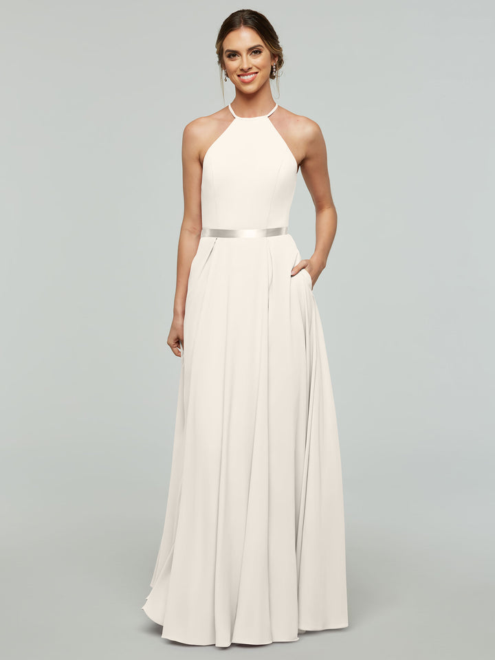 A woman models Avery Austin’s 80099—a sleeveless, white A-line dress with a halter neckline and satin ribbon belt. She smiles with light makeup, hair in a low bun, and poses with one hand in her pocket against a plain background.