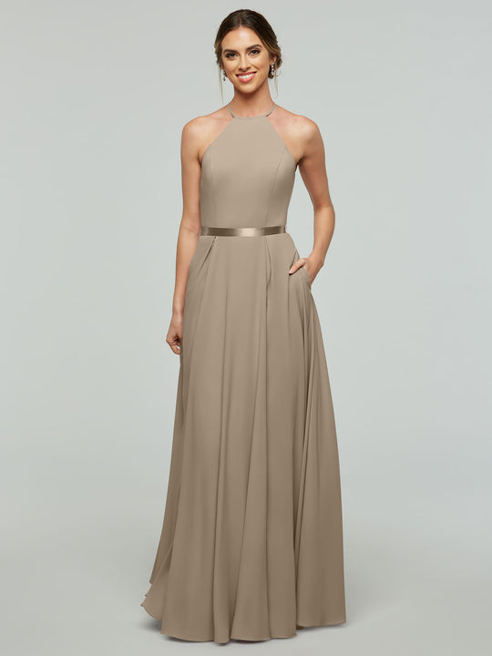 A woman faces forward, smiling with one hand in her pocket, wearing the Avery Austin 80099 dress— a sleeveless, floor-length taupe A-line style with a halter neckline and satin ribbon belt—against a plain light gray background.