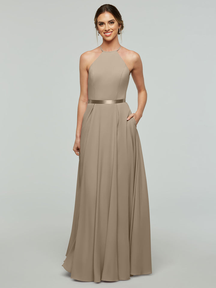 A woman faces forward, smiling with one hand in her pocket, wearing the Avery Austin 80099 dress— a sleeveless, floor-length taupe A-line style with a halter neckline and satin ribbon belt—against a plain light gray background.