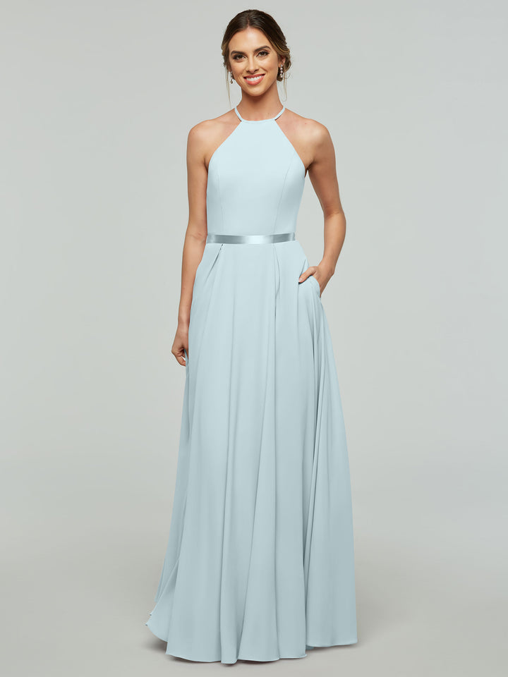 A woman smiles in the Avery Austin TH-80099 A-line sky blue dress, featuring a halter neckline, fitted bodice, satin ribbon belt, flowing skirt, and pockets. Her hair is styled back as she poses with one hand in her pocket.