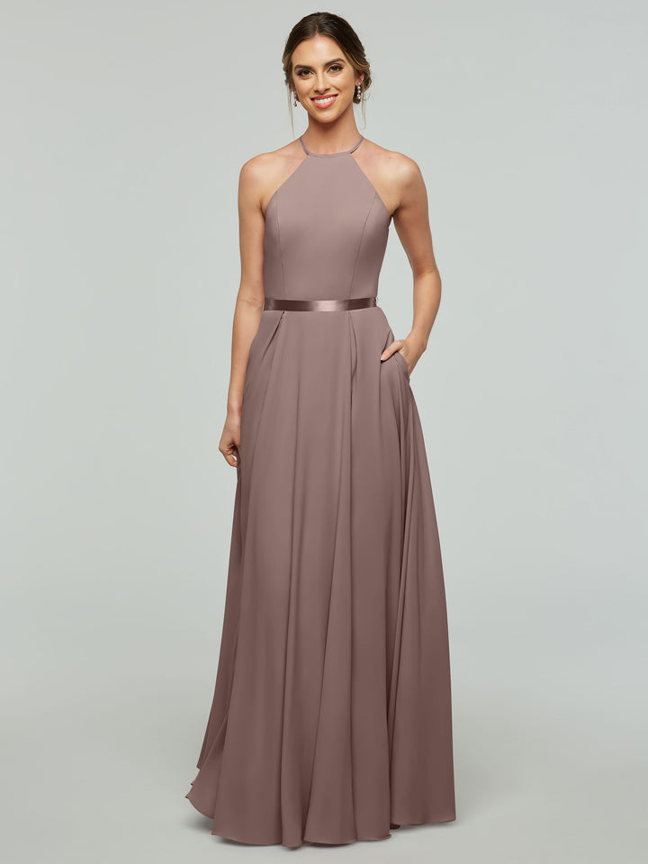 A woman wearing Avery Austin’s 80099, an A-line mauve gown with a halter neckline and satin ribbon belt, stands smiling with one hand in her pocket against a light gray background.