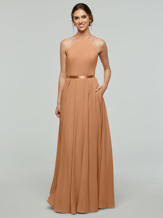 A woman smiles in Avery Austin’s 80099, a floor-length sleeveless A-Line dress with a halter neckline and satin waistband. Her hair is tied back, and with one hand in her pocket, she poses against a plain light background.