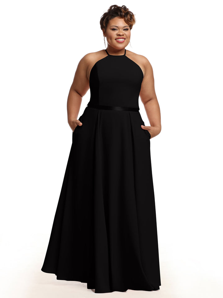A woman smiles with her hands in the pockets of Avery Austin’s 80099, a sleeveless black A-line gown with a fitted bodice and high halter neckline, standing against a white background.