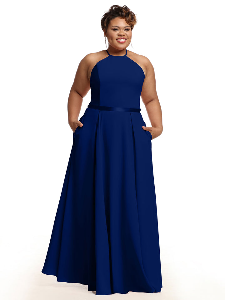 A woman smiles while standing with her hands in the pockets of Avery Austin’s 80099, a sleeveless floor-length dark blue A-line dress with a high halter neckline, against a plain white background.
