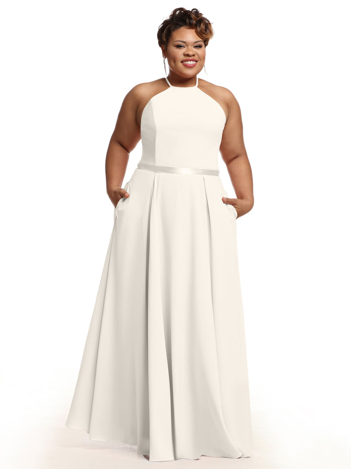A woman with short hair smiles while wearing Avery Austin’s 80099: a sleeveless, floor-length white A-line dress with a fitted bodice, halter neckline, and side pockets. She stands against a plain white background.