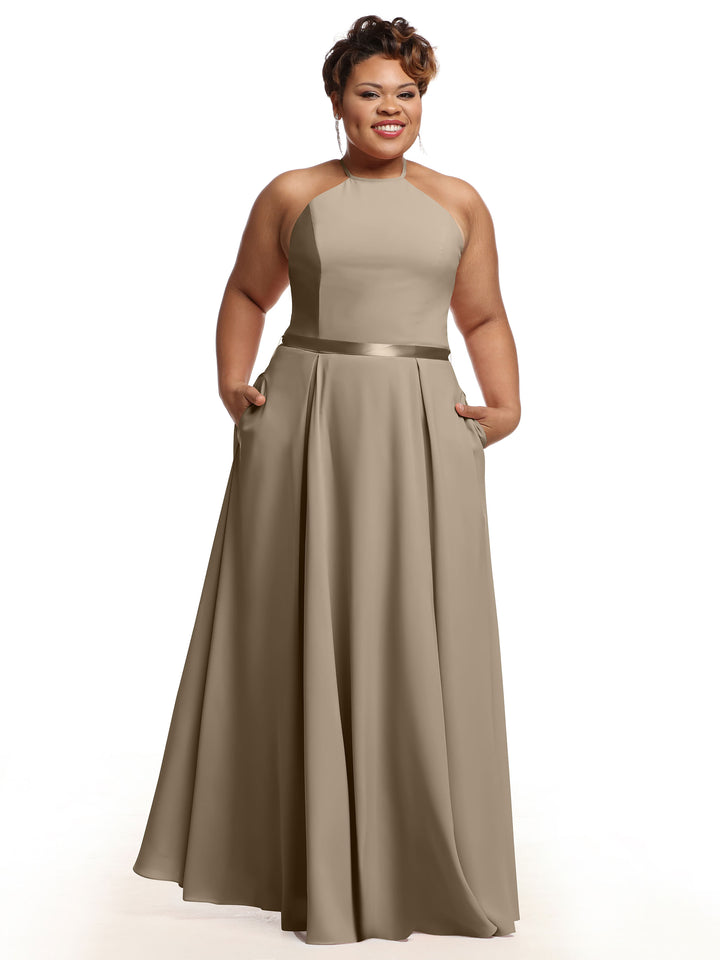 A woman stands smiling with her hands in the pockets of Avery Austin 80099, a sleeveless taupe A-line dress featuring a fitted bodice, halter neckline, and satin ribbon belt against a white background.