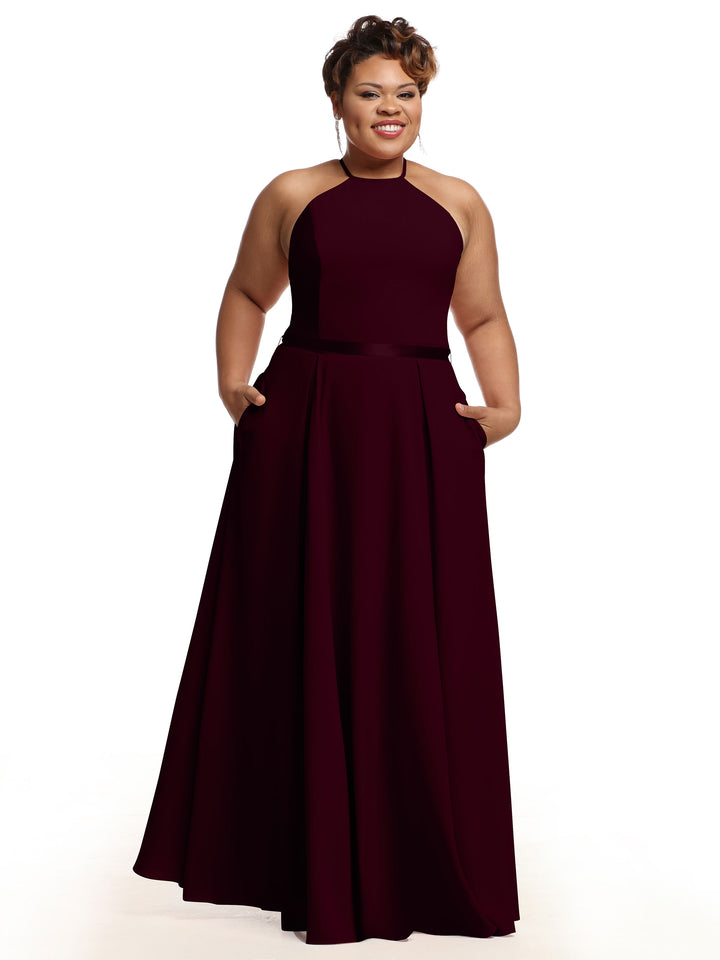 A woman stands smiling against a white background in Avery Austin 80099, a sleeveless, floor-length burgundy A-line dress featuring a high neckline and pockets.