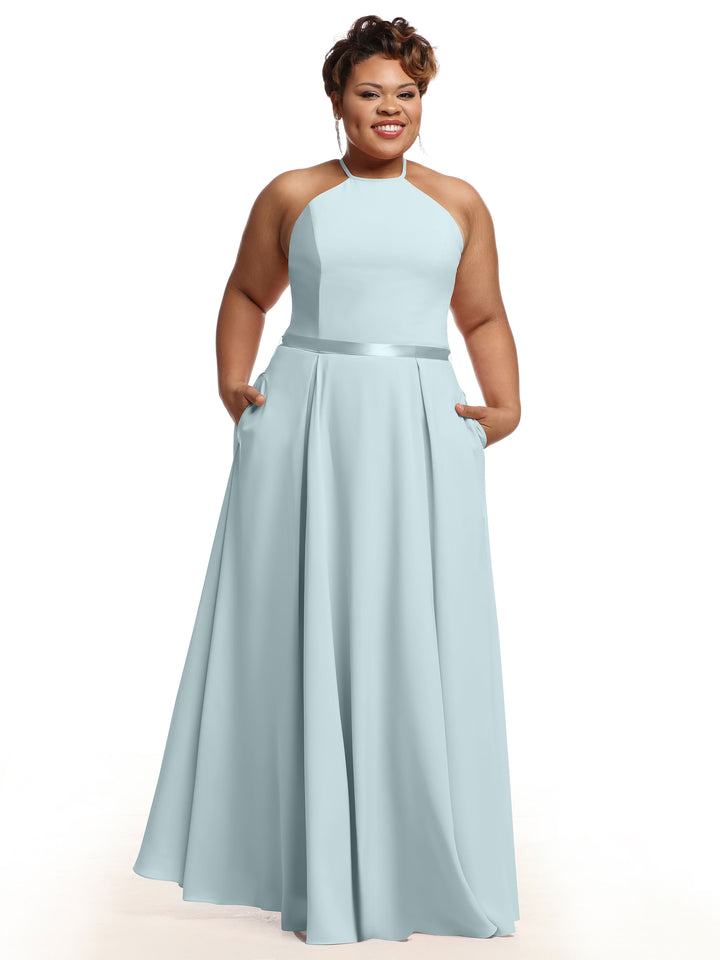 A woman stands and smiles against a white background, wearing Avery Austin’s 80099: a sleeveless light blue A-line dress with pockets and a satin ribbon belt.