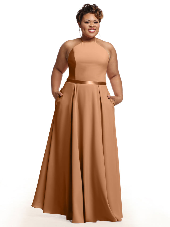 A woman smiles with her hands in her pockets, wearing Avery Austin’s sleeveless, floor-length A-Line dress (Style 80099) featuring a fitted bodice and satin ribbon belt, posed against a white background.