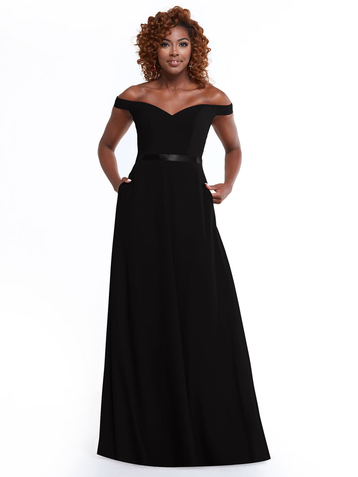 A woman with curly hair models Avery Austin’s 80100—a timeless, off-the-shoulder black gown with a fitted bodice and satin waistband—against a plain white background.