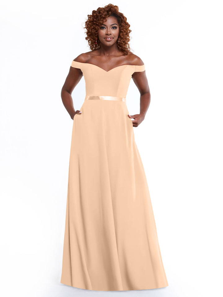 A woman with curly hair models Avery Austin’s 80100, a timeless peach off-the-shoulder A-line gown with a fitted bodice, satin waistband, and side pockets, as she stands gracefully against a white background.