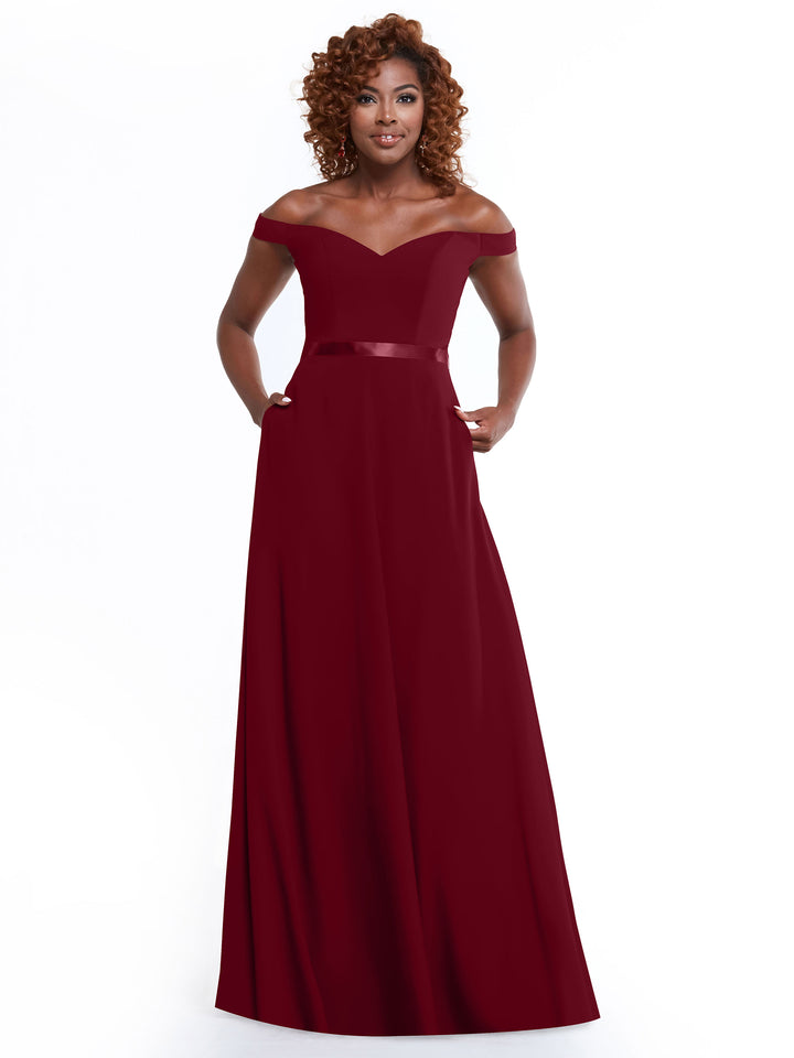 A woman with curly hair wears Avery Austin’s 80100: an off-the-shoulder, floor-length deep red A-line dress with a fitted bodice and satin waistband. She stands smiling against a white background, hands in her pockets.