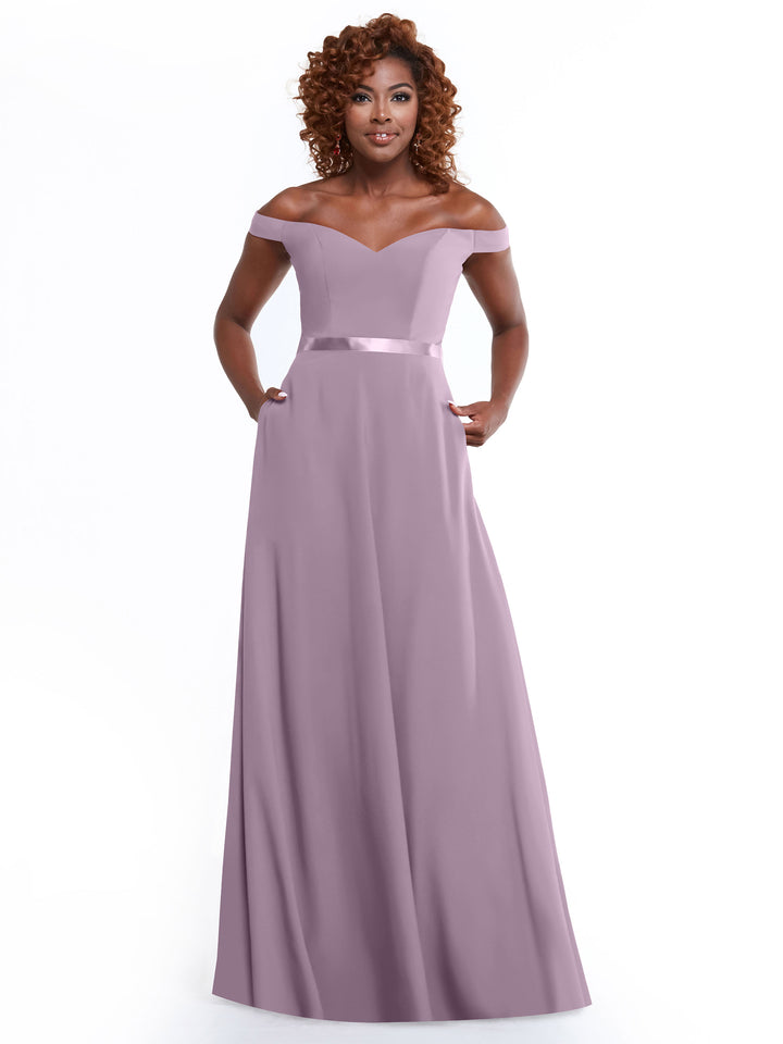 A woman smiles with her hands in the pockets of Avery Austin’s 80100—a lavender, off-the-shoulder, floor-length A-line dress featuring a satin waistband—against a plain white background.