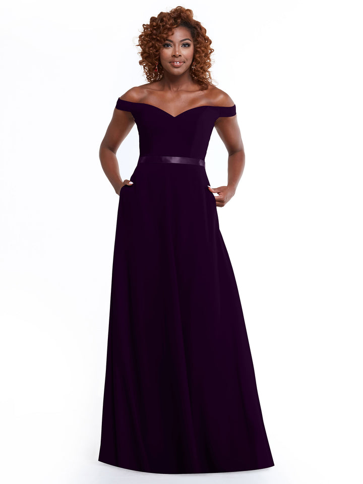 A woman with curly hair models Avery Austin’s 80100 dress, a classic dark purple off-the-shoulder style with a fitted bodice and subtle sheen at the waist, against a plain white background.