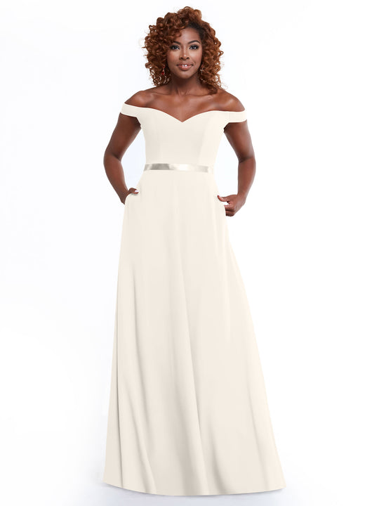 A woman with curly hair models Avery Austin’s 80100: a timeless, off-the-shoulder A-Line dress with a fitted bodice, satin waistband, and pockets. She stands confidently against a white background.