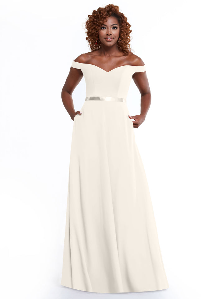 A woman with curly hair models Avery Austin’s 80100: a timeless, off-the-shoulder A-Line dress with a fitted bodice, satin waistband, and pockets. She stands confidently against a white background.