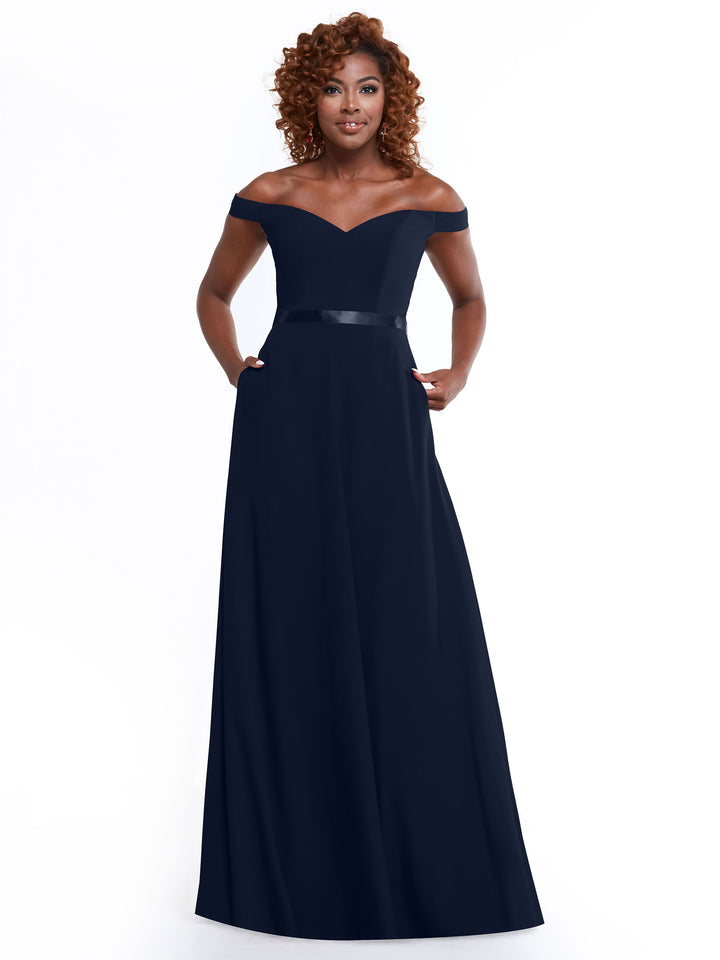 A woman with curly hair confidently poses with hands in pockets, wearing Avery Austin’s timeless 80100—an elegant, floor-length, off-the-shoulder navy blue gown with a fitted bodice and subtle ribbon belt—against a white background.