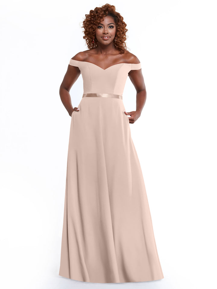 Woman wearing Avery Austin 80100, an off-the-shoulder A-line dress in rose pink with a satin waistband and side pockets, standing and smiling against a plain white background.